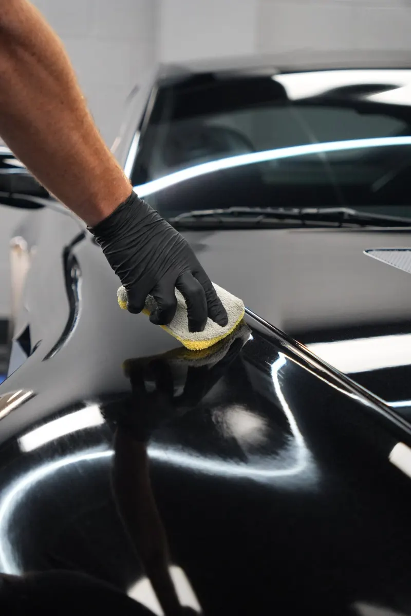 A black-gloved hand presses a foam applicator pad onto the curved black fender of a car, with light strips reflected across the panel.
