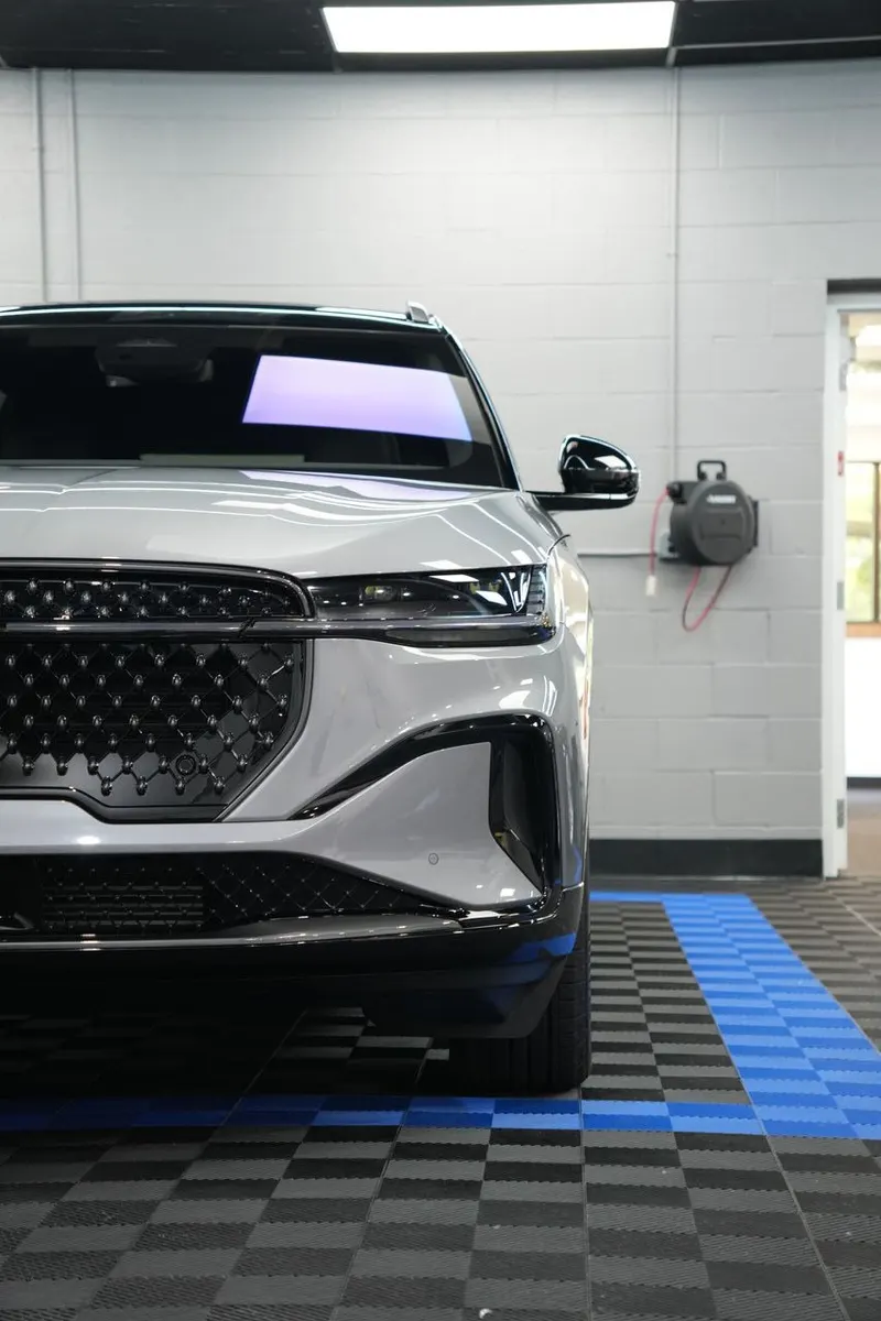 Front end of a vehicle parked in a shop bay, with equipment on the floor and a garage door in the background.
