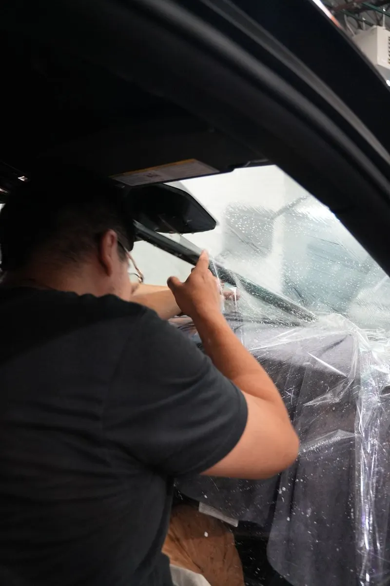 A technician applies film to a vehicle's rear window from inside the cabin, with seats covered in plastic.