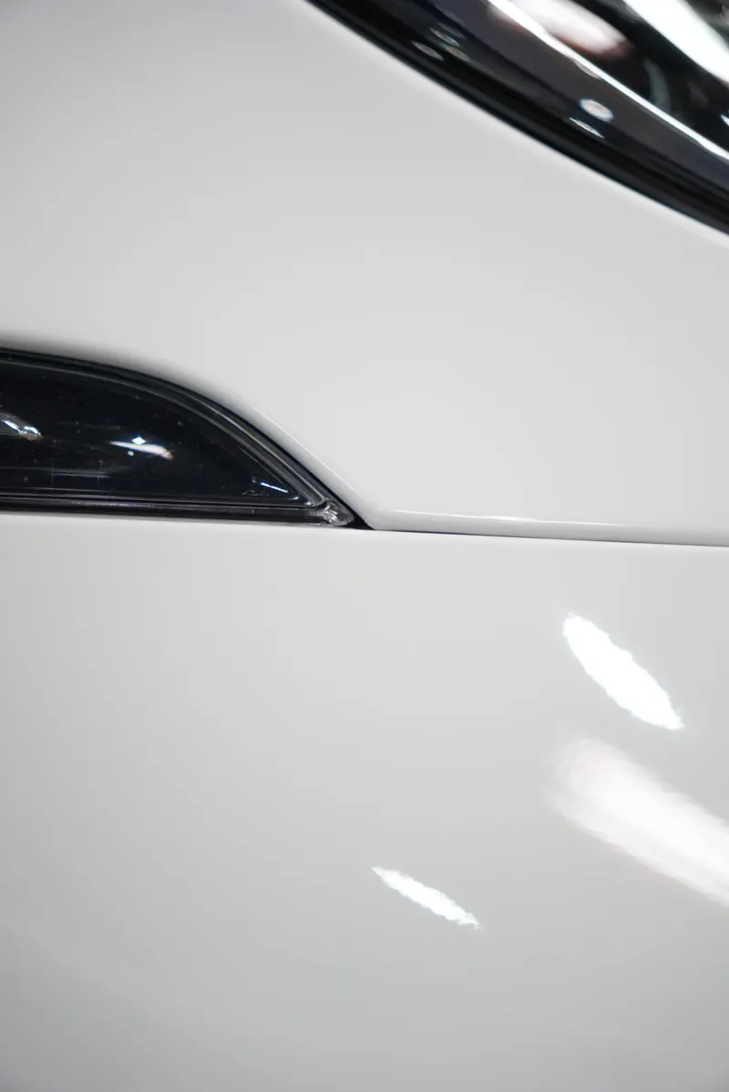 Close-up of a white glossy fender meeting a dark headlight edge, with a diagonal panel shutline and reflections of shop lights.