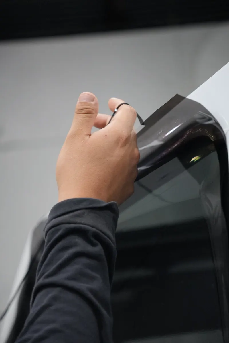 A hand holds a strip of dark window film at the upper edge of a vehicle's window, beside the car's white body.