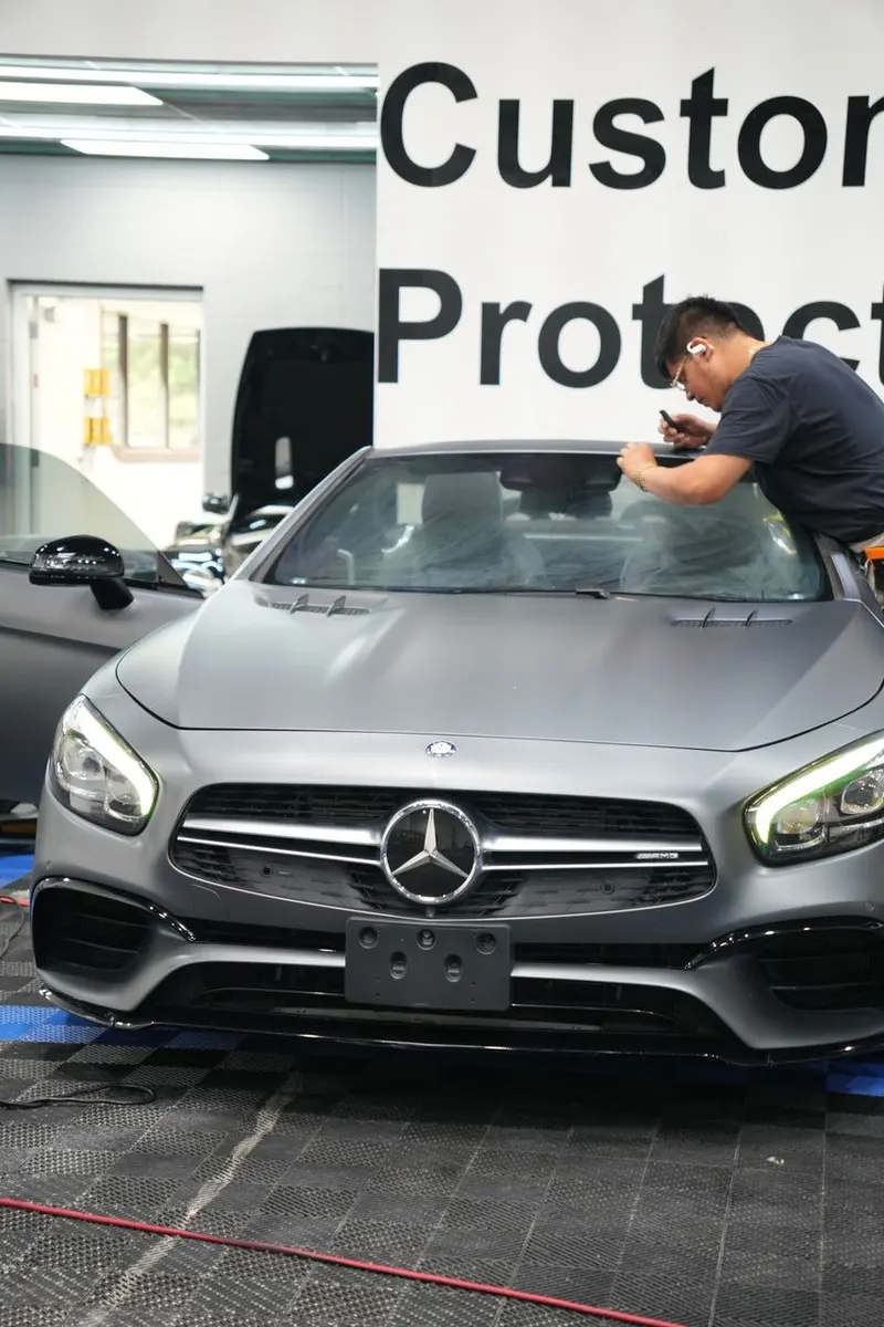 A technician applies film to a vehicle's windshield in a shop bay, with wall signage in the background.